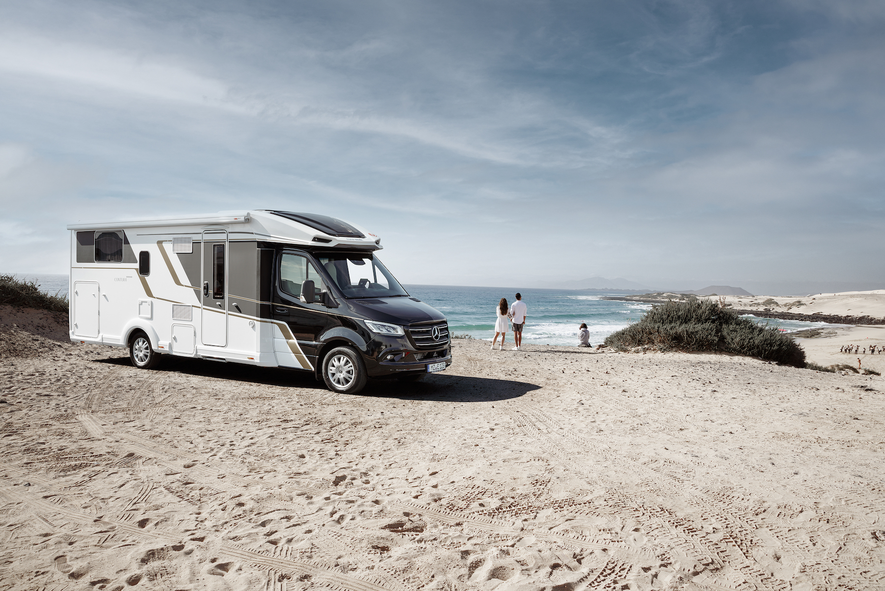 Wohnmobil am Strand, daneben einige Personen und Meer im Hintergrund.