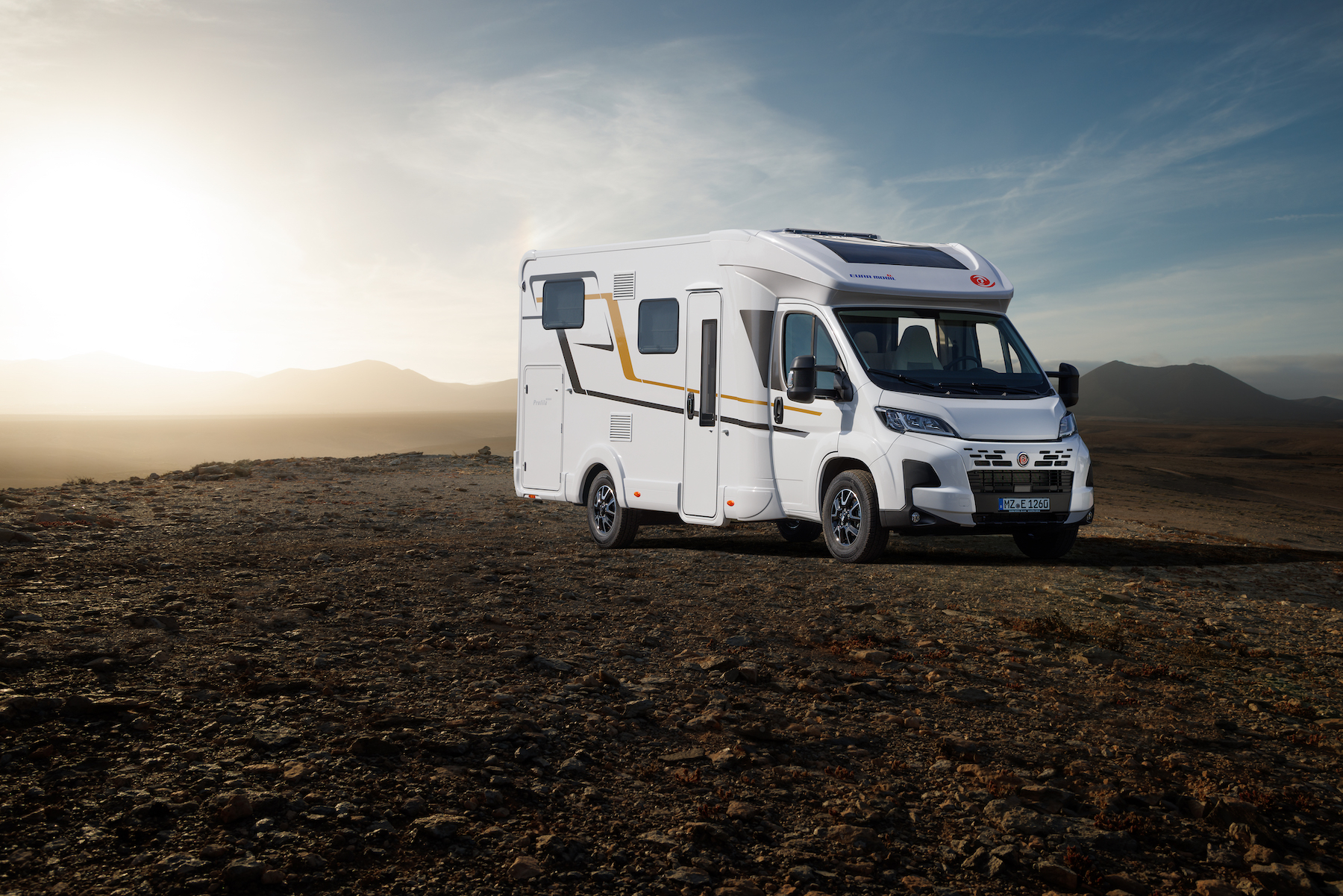 Auf Wiese stehendes Wohnmobil, Umrisse der Landschaft im Hintergrund beleuchtet von untergehenden Sonne.